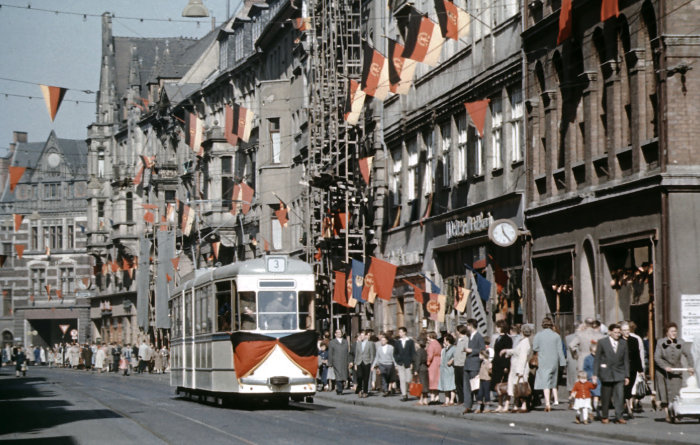 Eine mit zahlreichen DDR-, FDJ- und Arbeiterfahnen geschmückte Häuserzeile. Im Vordergrund eine mit einer Fahne geschmückte Straßenbahn.