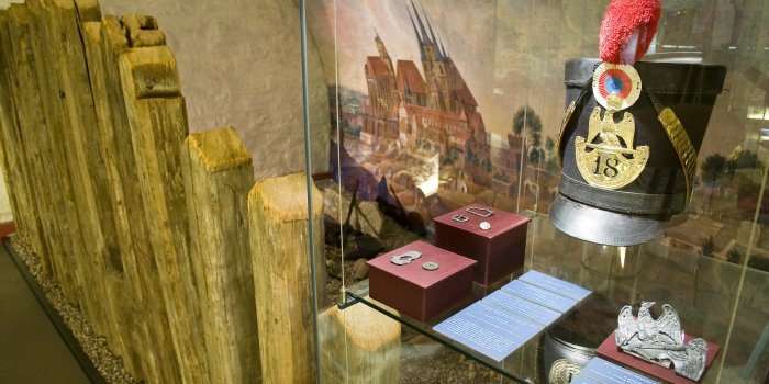Links ein Staketenzaun, rechts eine Vitrine mit verschiedenen Ausstellungsstücken.