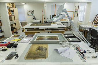 Werkstattraum mit verschiedenen Arbeitstischen, auf denen Gemälde und Werkzeuge liegen