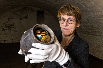 eine Frau trägt weiße Handschuhe und hält einen Krug mit historischen Münzen