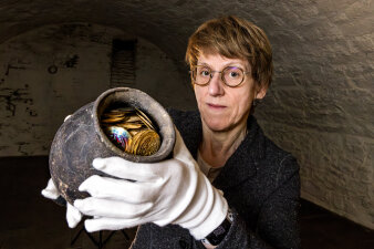 eine Frau trägt weiße Handschuhe und hält einen Krug mit historischen Münzen