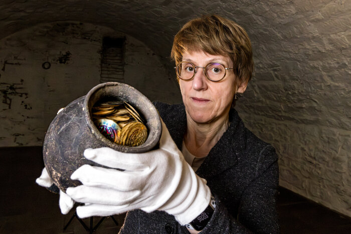 eine Frau trägt weiße Handschuhe und hält einen Krug mit historischen Münzen