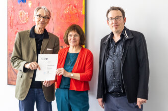 ein Mann und eine Frau halten gemeinsam ein bedrucktes Blatt Papier in der Hand, rechts daneben steht ein weiterer 