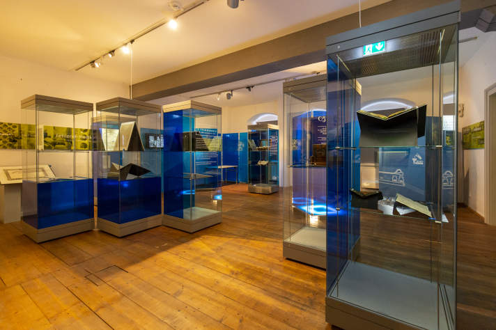 Ausstellungsraum mit hohen Vitrinen in blau mit Aufschriften in weiß und Exponaten wie Bildern.