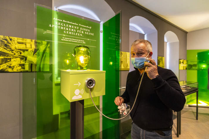 Ein Mann bedient zwischen grünen Vitrinen einen Audioguide in einem Museum, schaut in die Kamera.