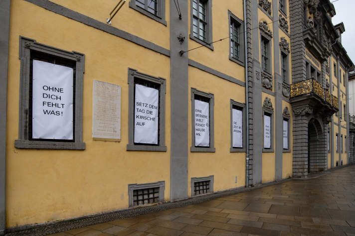 Aufgehängte weiße Banner mit schwarzer Schrift in Fenstern eines gelben Gebäudes
