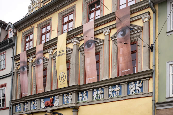Banner mit Augen und Gesichtern an Fassade eines gelben verzierten Gebäudes