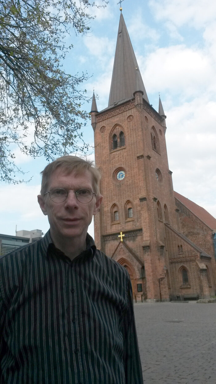 Mann mit Brille und schwarzen Hemd, vor einer kleinen Kirche