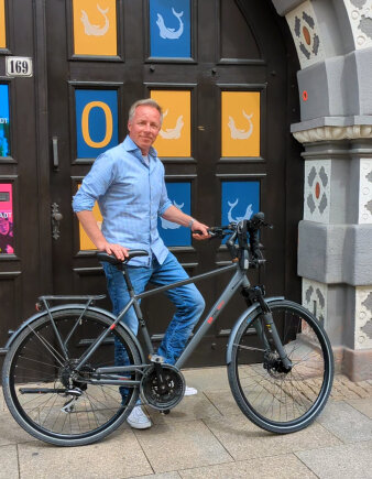 Mann mittleren Alters im Hemd steht mit einem Fahrrad vor einem großen, bunten Holztor
