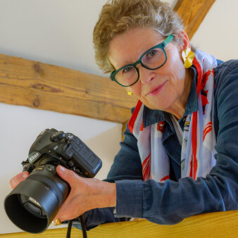 Frau mittleren Alters mit grüner Brille und Kamera in der Hand
