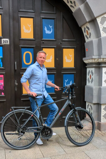 Mann mittleren Alters im Hemd steht mit einem Fahrrad vor einem großen, bunten Holztor