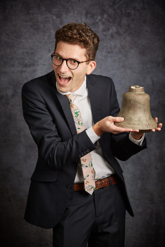 Junger Mann im Anzug mit Brille hält eine Glocke in der Hand