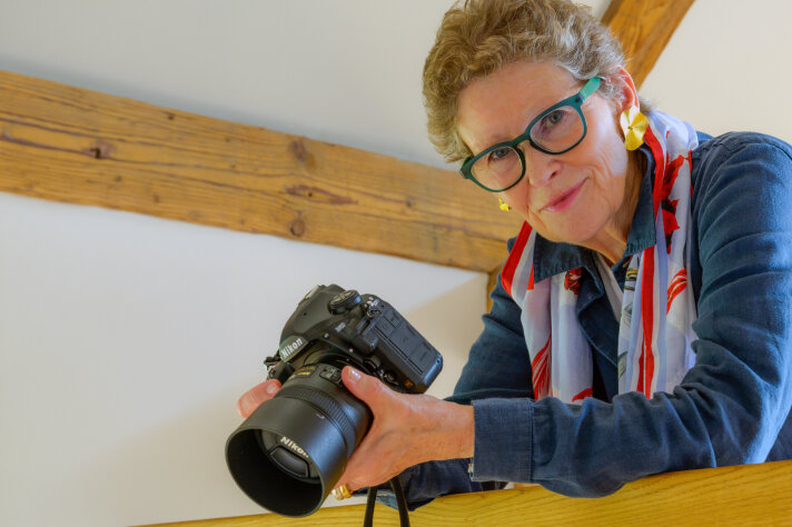 Frau mittleren Alters mit grüner Brille und Kamera in der Hand