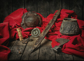 Metallener Helm, Waffen und Werkzeuge auf urigem Holztisch umringt von rotem Samtstoff vor dunklem Hintergrund