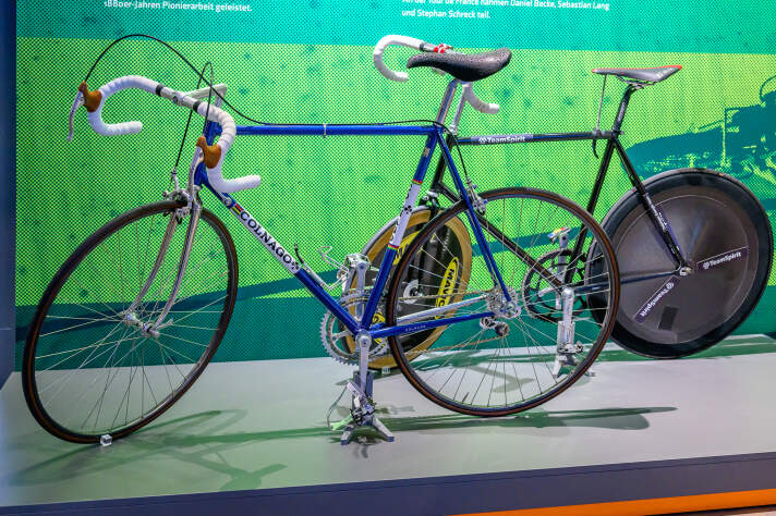 Im Vordergrund ein blaues modernes Rennrad und im Hintergrund ein schwarzes Bahnrennrad ohne Bremsen und Schaltung.