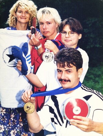 Eine Frau hält ein T-Shirt hoch und vor ihr befinden sich drei Sportler und halten ihre Medaillen hoch.