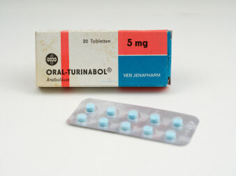Eine weiß-rot-blaue Faltschachtel mit Aufdruck „Oral-Turinabol“ 5 mg, VEB Jenapharm, davor die blaue Tabletten.