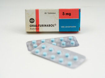 Eine weiß-rot-blaue Faltschachtel mit Aufdruck „Oral-Turinabol“ 5 mg, VEB Jenapharm, davor die blaue Tabletten.