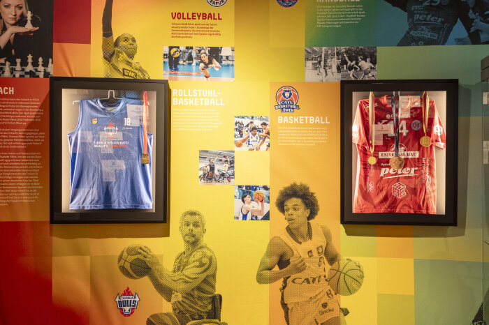 bunte Aussstellungswand mit Fotos und zwei gerahmten Basketball-Trikots