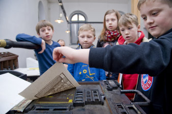 Kinder stehen an einer Druckerpresse und drucken eine Buchseite