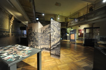 Raum mit einer Ausstellung. Im Vordergrund eine Ausstellungstafel mit einer mittelalterlichen Stadtansicht, davor ein Tisch, auf dem man die Ansicht aus Puzzleteilen zusammensetzen kann