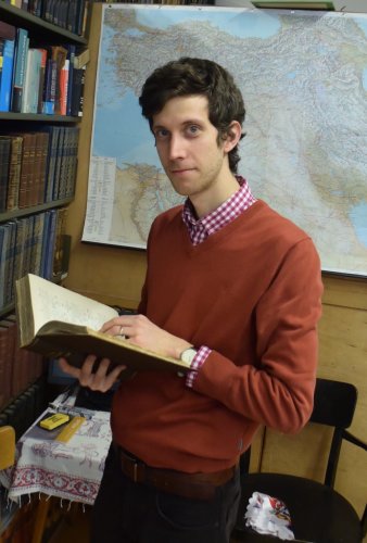 Ein Mann dunklen Haaren steht vor einer Landkarte. Er hält ein Buch in der Hand und blickt in die Kamera. 
