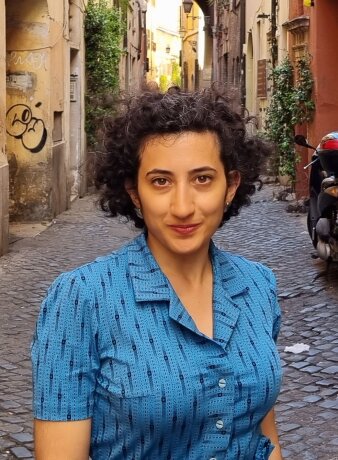 Portrait einer Frau mit schwarzen Haaren und einem blauen Oberteil in einer Gasse mit Häusern im Hintergrund