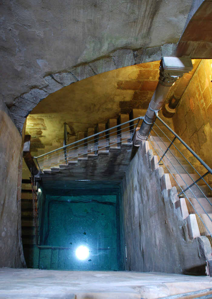 Ein dunkler Raum mit einer abfallenden Treppe und einem Lichtpunkt im Wasser in der Tiefe