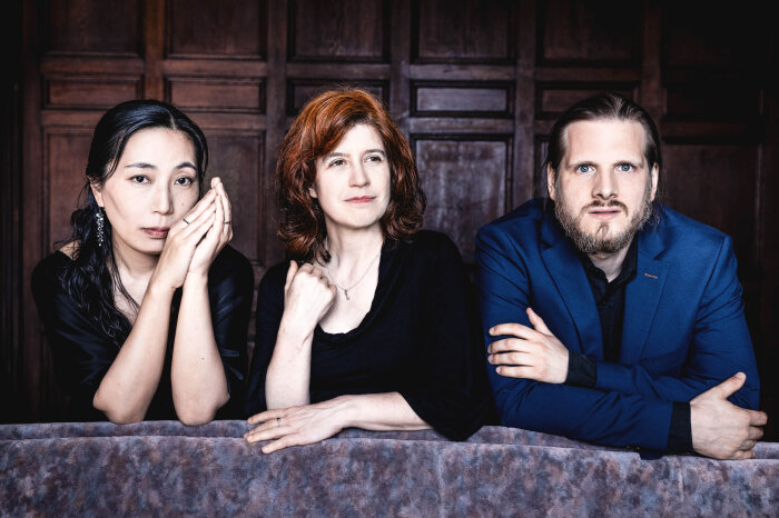 Zwei Frauen und ein Mann mittleren Alters in dunkler Kleidung vor einer dunkelbraunen Holzwand sitzend
