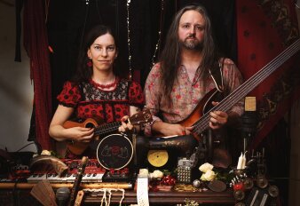 Eine Frau und ein Mann mit Saiteninstrumenten in der Hand hinter einem Tisch mit weiteren Instrumenten stehend.