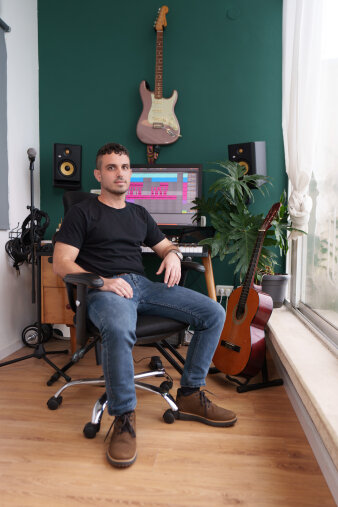 Junger Mann auf einem Stuhl sitzend in einem Musikstudio, umringt von Instrumenten, einem Mikrofon und PC
