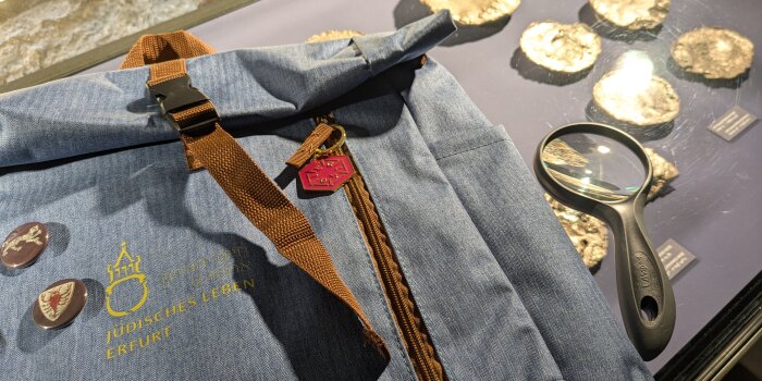 Ein kleiner, graublauer Rucksack mit Lupe und Buttons liegt auf einer Museumsvitrine mit gold-glänzenden Exponaten