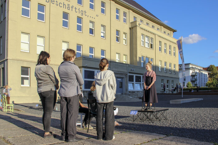 eine Person steht auf einer kleinen Bühne, weitere Personen hören zu