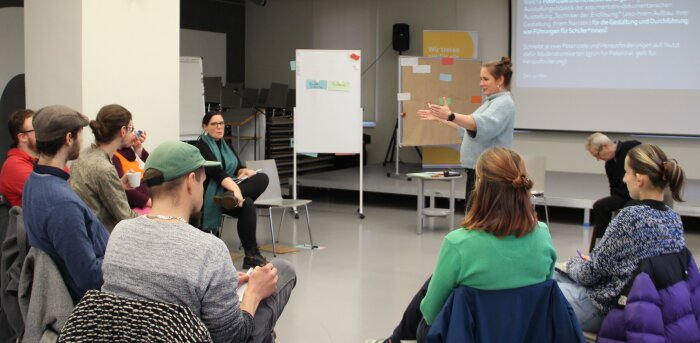 Im Seminarraum steht eine Moderatorin und erklärt etwas, während eine Gruppe von Teilnehmenden im Stuhkreis sitzt