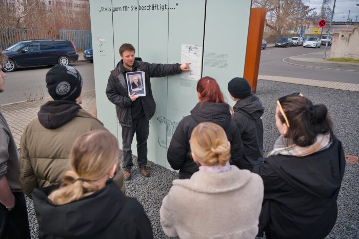 Gruppe von Menschen steht vor Stele einer Außenausstellung während Guide auf ein Dokument zeigt.
