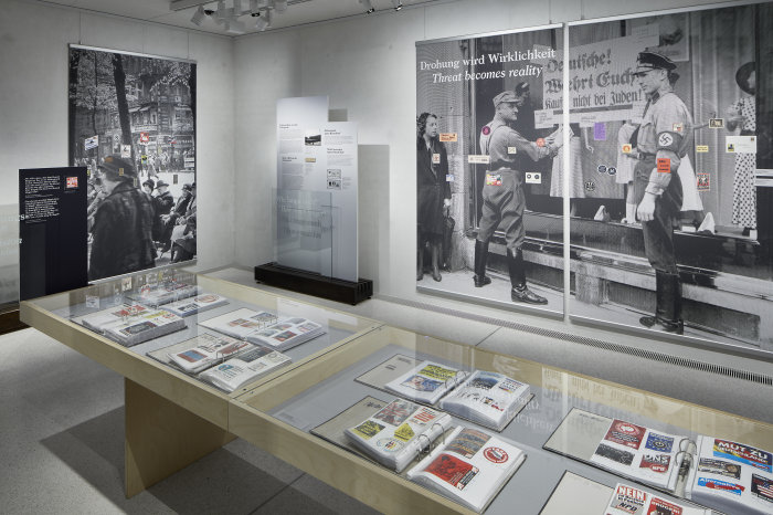 Blick in die Ausstellung "Angezettelt. Antisemitische Aufkleber von 1880 bis heute" Ausstellungstafeln und Vitrinen der Ausstellung sind zu sehen. Auf einer Tafel steht "Drohung wird Wirklichkeit".