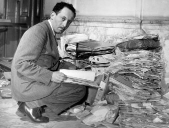 schwarzweiß Foto von Mann der vor einem Berg von Unterlagen hockt
