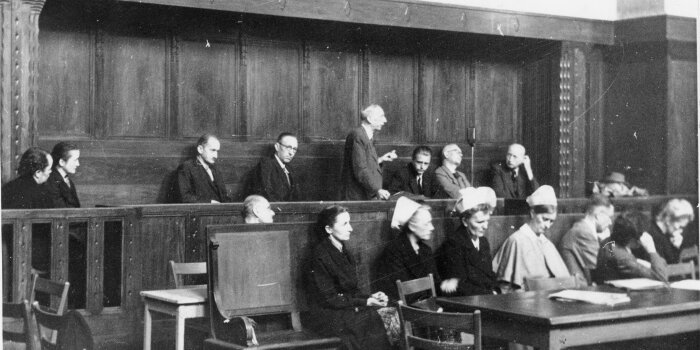 Schwarzweiß Fotografie. Frauen und Männer auf einer hölzernen Anklagebank in einem Gerichtsraum.